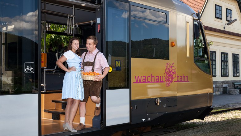 Mit der Wachaubahn zum Spitzer Marillenkirtag, © NÖVOG/citronenrot Pärchen in Trachtenkleidung mit einem Korb voll Marillen steht beim Eingang der Wachaubahn