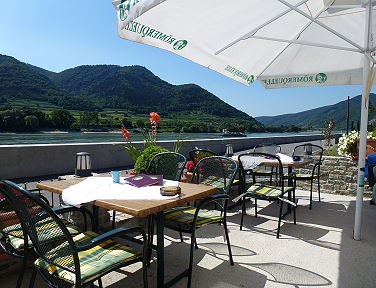 Zahrada pro hosty s výhledem na Dunaj, © Fam. Pichler Zahrada pro hosty se stoly a židlemi pod slunečníky s výhledem na Dunaj a zalesněné kopce v pozadí.