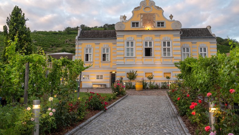 Domäne Wachau, Kellerschlössl Dürnstein, © Domäne Wachau Gelbes Barockgebäude mit Garten und Weinreben, Dämmerung.