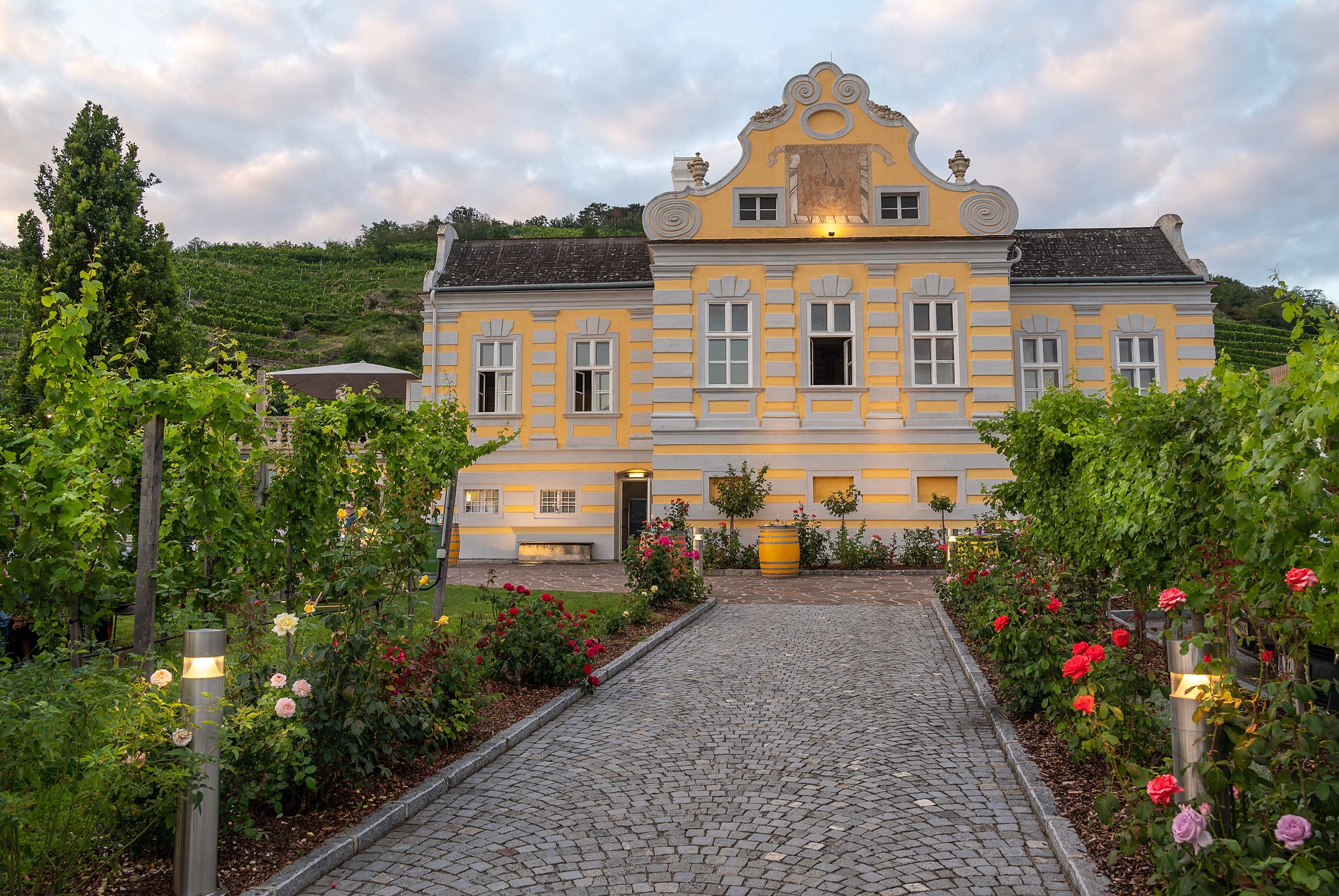 Gelbes Barockgebäude mit Garten und Weinreben, Dämmerung.