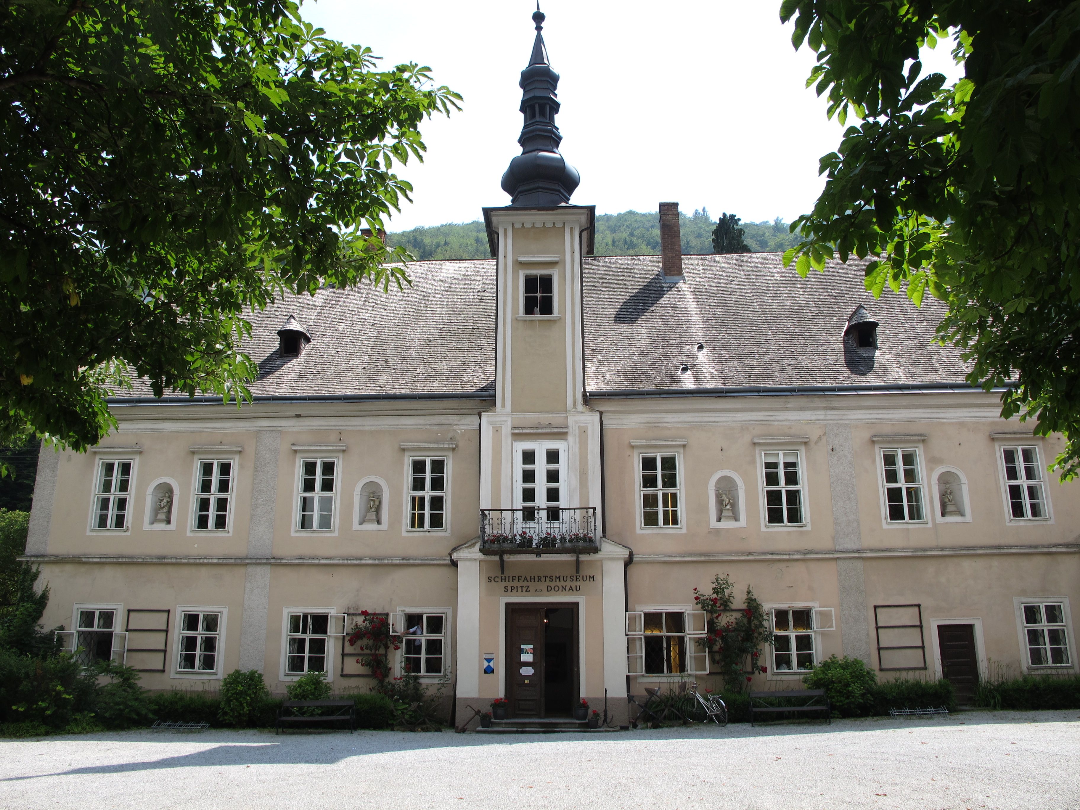 Außenansicht eines historischen Gebäudes mit Schindeldach und Turm, umgeben von Bäumen.