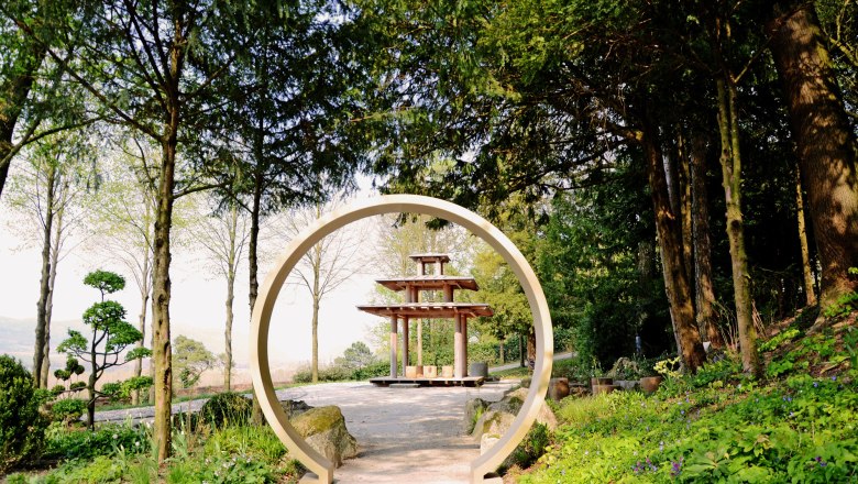 Ein orientalischer Garten mit einem runden Torbogen und einer Pagode im Hintergrund, umgeben von B&auml;umen und Pflanzen.