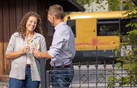 Mann und Frau sto&szlig;en mit Weinglas an. Im Hintergrund steht die Wachaubahn