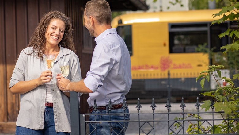 Mann und Frau sto&szlig;en mit Weinglas an. Im Hintergrund steht die Wachaubahn