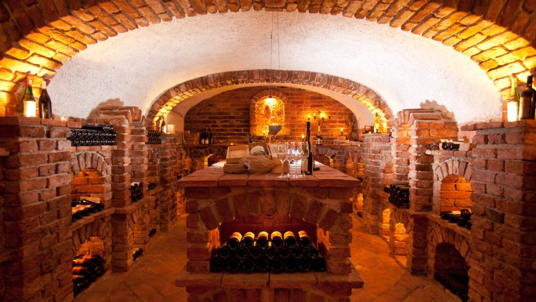 Ein stimmungsvoll beleuchteter Weinkeller mit Backsteinw&auml;nden und Weinflaschen in Regalen.