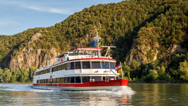 Ein Passagierschiff f&auml;hrt auf einem Fluss vor einer bewaldeten H&uuml;gelkulisse.