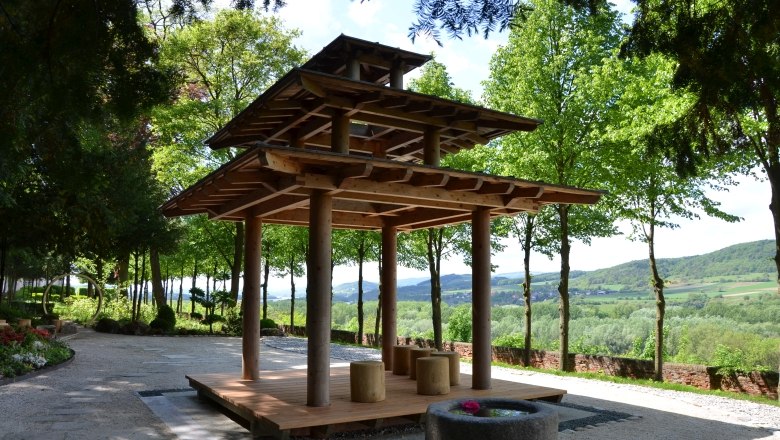 Asiatischer Holzpavillon in einem Garten mit Bäumen und Landschaft im Hintergrund.