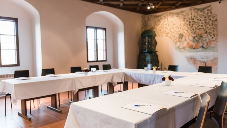 Tagungsraum mit U-förmiger Tischanordnung, weißen Tischdecken und Schreibmaterialien. Im Hintergrund ein Wandgemälde und ein Kachelofen.