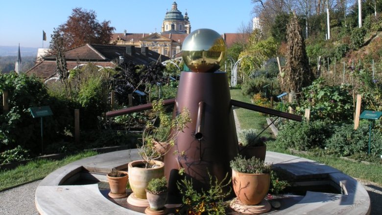 Ein dekorativer Garten mit einer Skulptur im Vordergrund und einem historischen Gebäude im Hintergrund.
