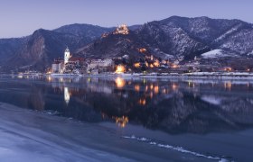 Winterliche Ansicht von D&uuml;rnstein an der Donau mit beleuchteten Geb&auml;uden und schneebedeckten H&uuml;geln im Hintergrund.