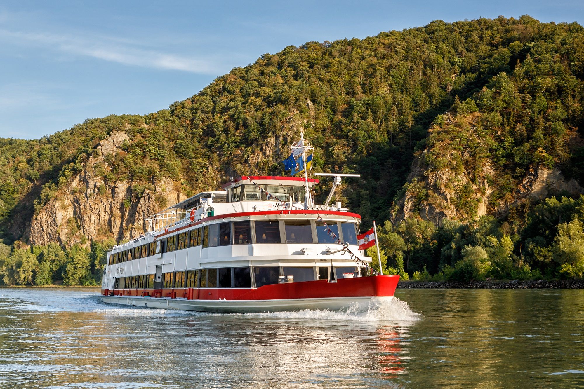 Ein Passagierschiff fährt auf einem Fluss vor einer bewaldeten Hügelkulisse.