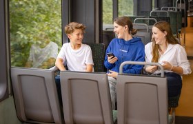 Bursche und 2 Mädchen im Zug. Die Mädchen haben Mobiltelefone in der Hand und Kopfhöhrer im Ohr.