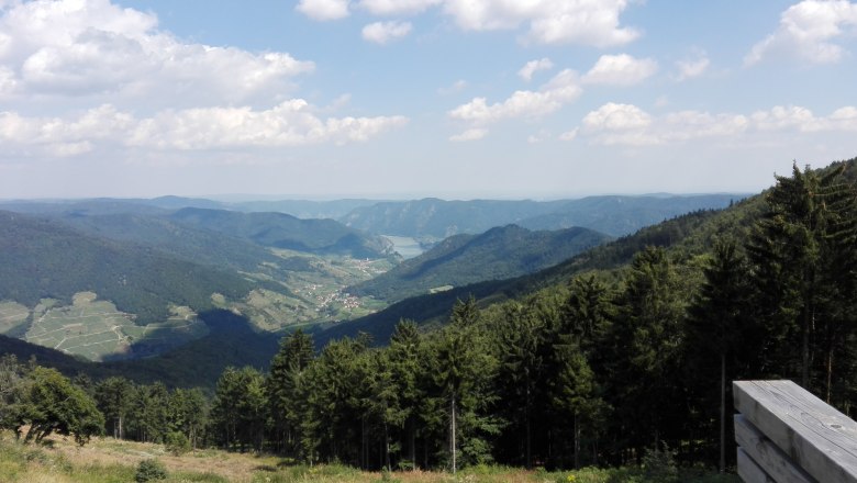 Panoramablick &uuml;ber bewaldete H&uuml;gel und ein Tal im Naturpark Jauerling, mit blauem Himmel und Wolken.