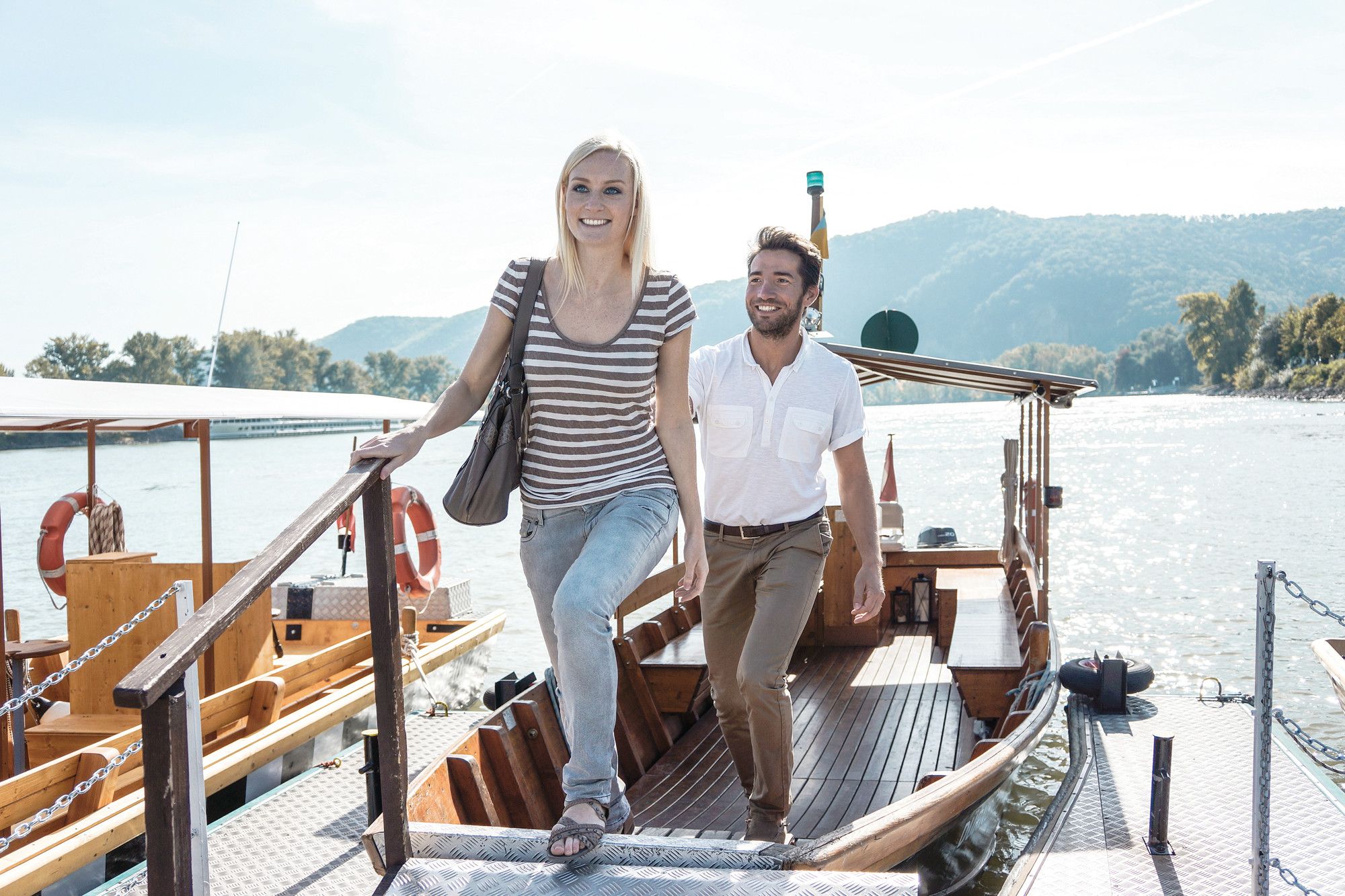 Ein Mann und eine Frau steigen von einem Boot auf einen Steg. Im Hintergrund ist ein Fluss und bewaldete Hügel zu sehen.