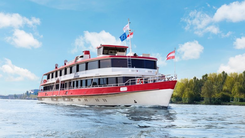 Ein Passagierschiff mit roten und wei&szlig;en Farben f&auml;hrt auf einem Fluss unter blauem Himmel.