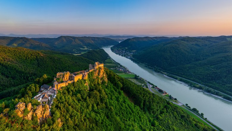 Luftaufnahme der Burgruine Aggstein an der Donau, umgeben von grünen Hügeln und Wäldern bei Sonnenuntergang.