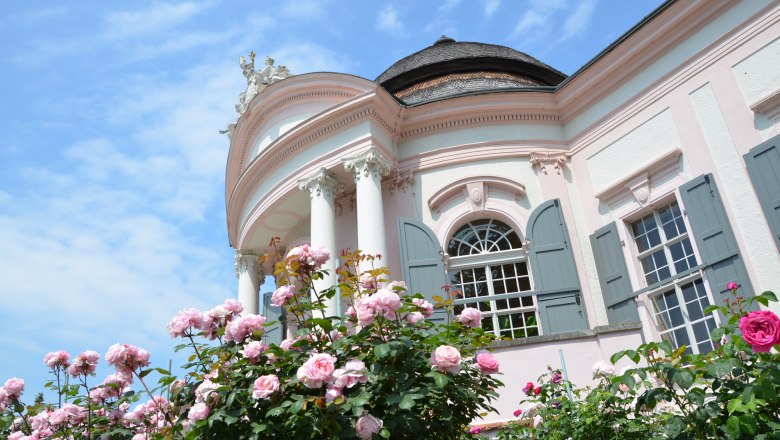 Barocker Pavillon mit rosa Fassade und bl&uuml;henden Rosen im Vordergrund.